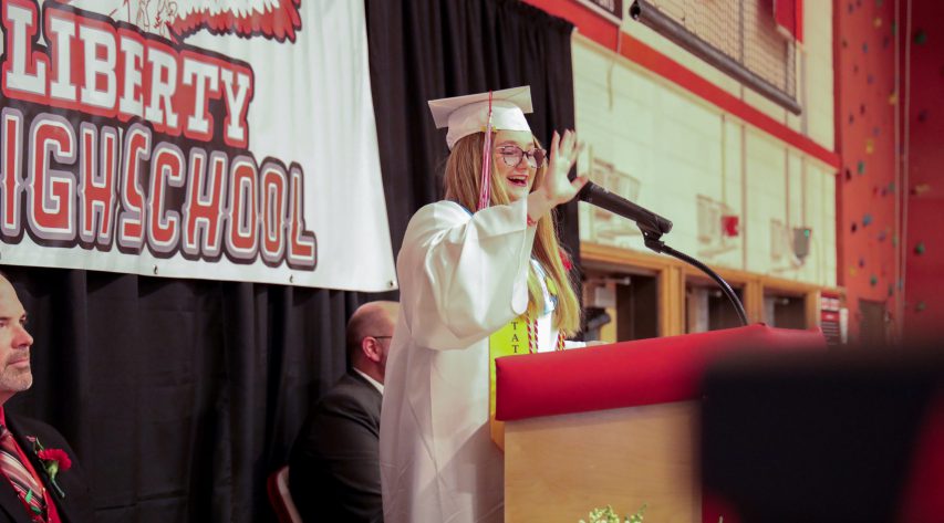 A student in a white cap and gown waves from the podium