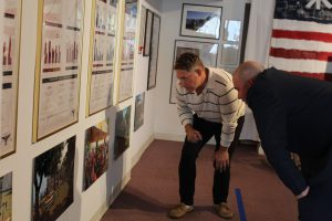 Two men examine framed charts and photographs displayed on a wall, with an American flag in the background.