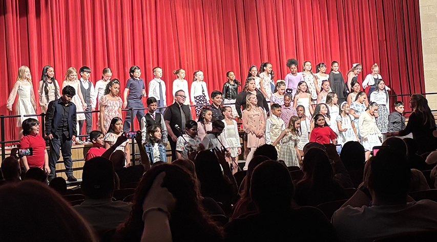 Elementary students sing on risers in front of a stage with red curtains