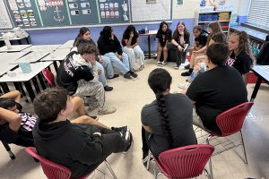 A diverse group of teenagers sits in a circle in a classroom, engaged in discussion.