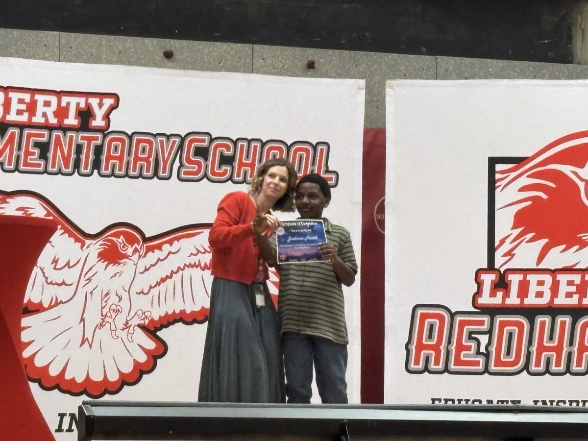 A woman in a red cardigan presents a certificate to a young boy in a striped shirt on a stage.