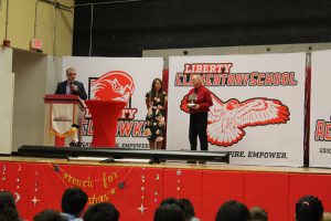 A man holds a tree award as two others watch in front of a Liberty Elementary sign