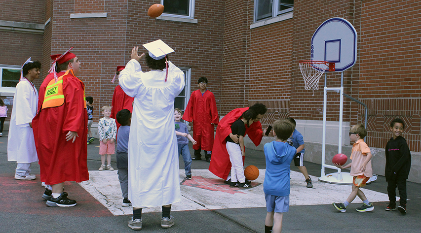 A mix of seniors in cap and gowns and younger children play with footballs and basketballs on a pavement court