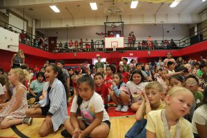 A large group of children sit on a gymnasium floor, watching an event.