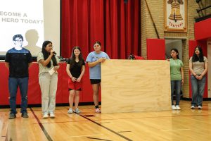 Students stand on a gymnasium floor in front of a large wooden panel and red curtains.