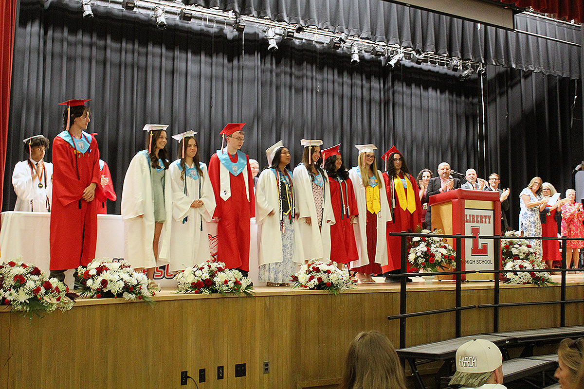 Graduates in red and white gowns stand on a stage with a black curtain backdrop.