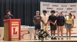 A speaker stands at a podium with the Liberty High School logo, addressing a group of students on stage.