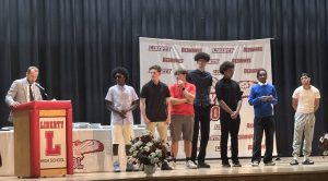 A man in a suit speaks at a podium with the Liberty High School logo, while seven young men stand on stage.