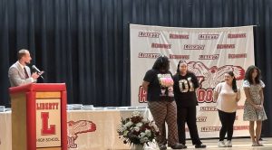 A man in a suit speaks at a podium with "Liberty High School" on it, while four students stand on stage.