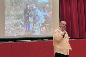 A man with facial scars speaks into a microphone in front of a projection screen showing a military-style photo.