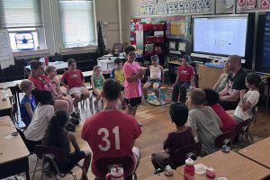 A diverse group of children and adults are seated in a circle in a classroom, facing a boy in a pink shirt.