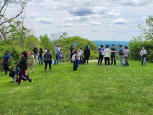 A diverse group of people stand on a grassy hillside with a scenic view of rolling hills and a cloudy sky.