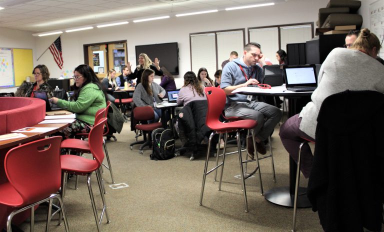 Teachers sit in groups at tables as a person speaks