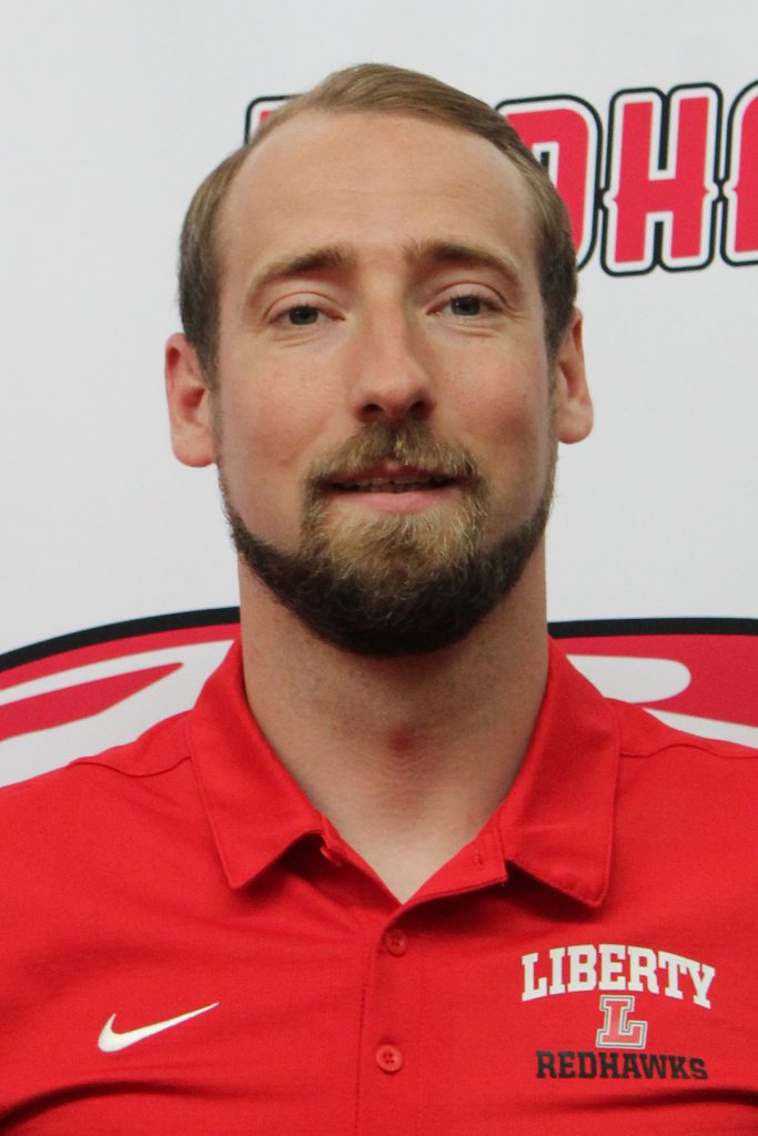 Headshot of thomas zehner, a smiling man with a beard and short brown hair wearing a red Liberty Redhawks polo shirt.