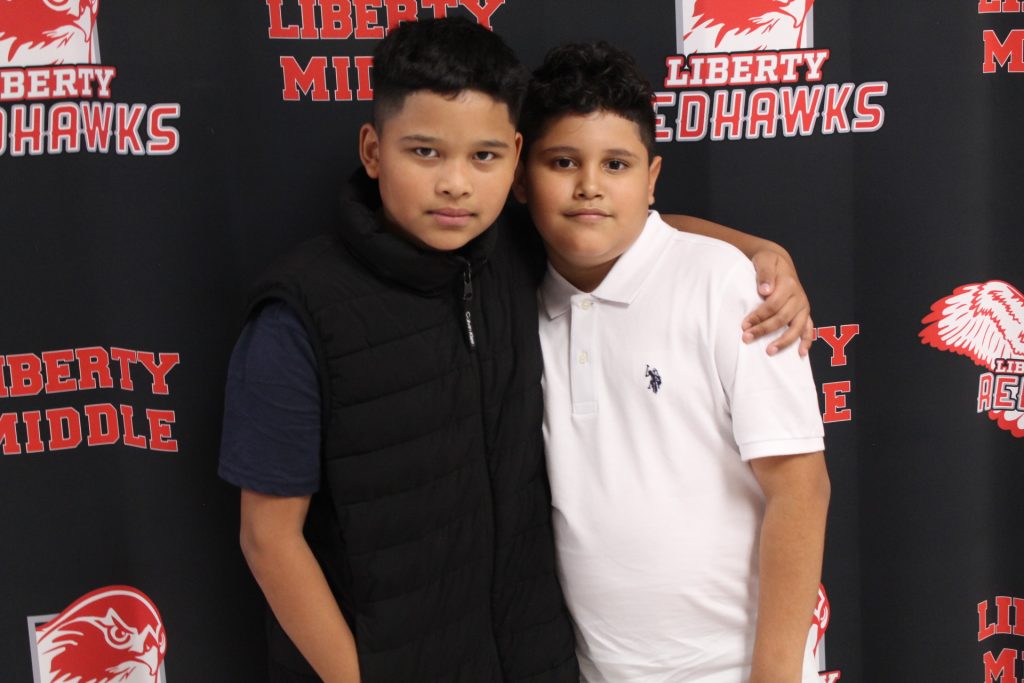 two middle school students posing for a photo, standing against a liberty middle school background with the liberty red hawks logo, arms around each other