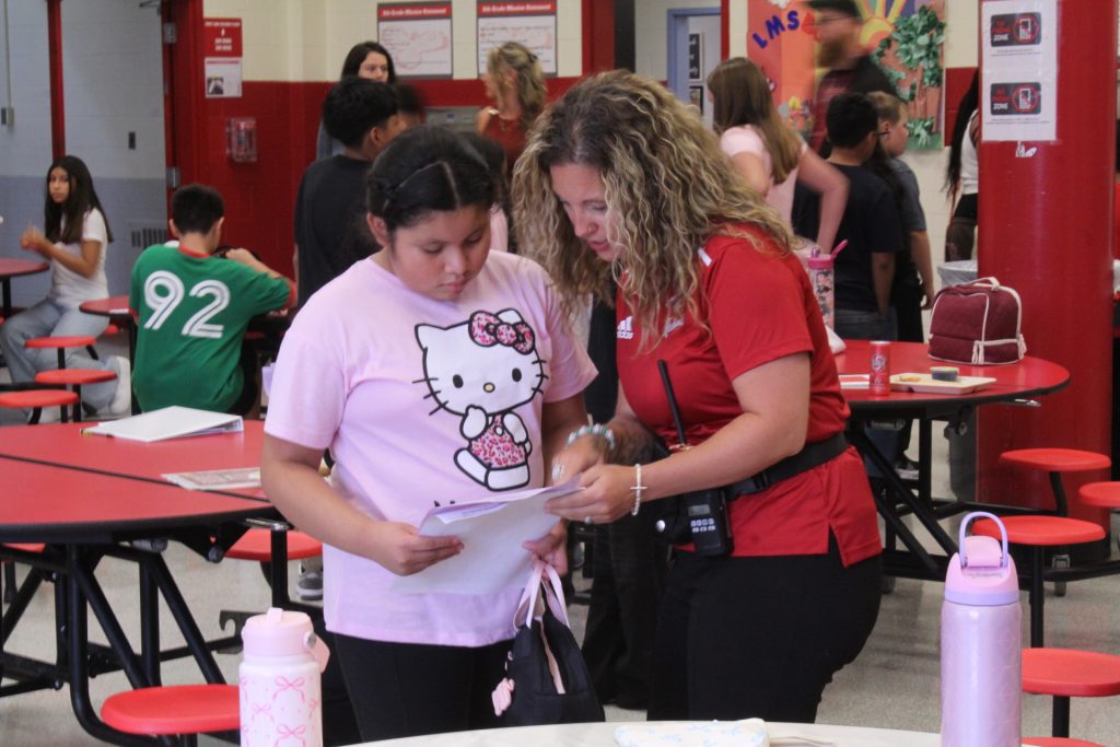 inside the middle school lunch room, a student is being assisted by a teacher, they are both looking at a paper