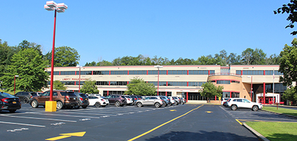 libert middle school, landscape view of building, tan coloured siding, dark windows with red panes, cars out front in parking lot with large standing light with red pole and yellow cement holding it up